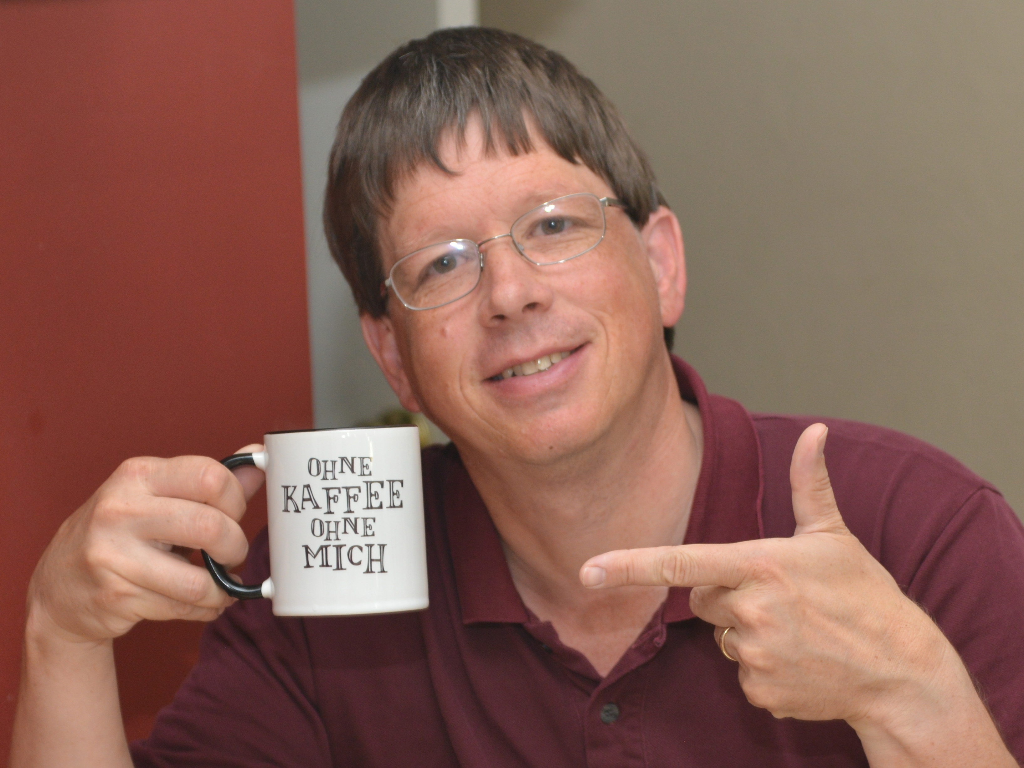 Gerd looks into the camera smiling, pointing to his coffee cup. On his cup are the words "without coffee, without me" in German.