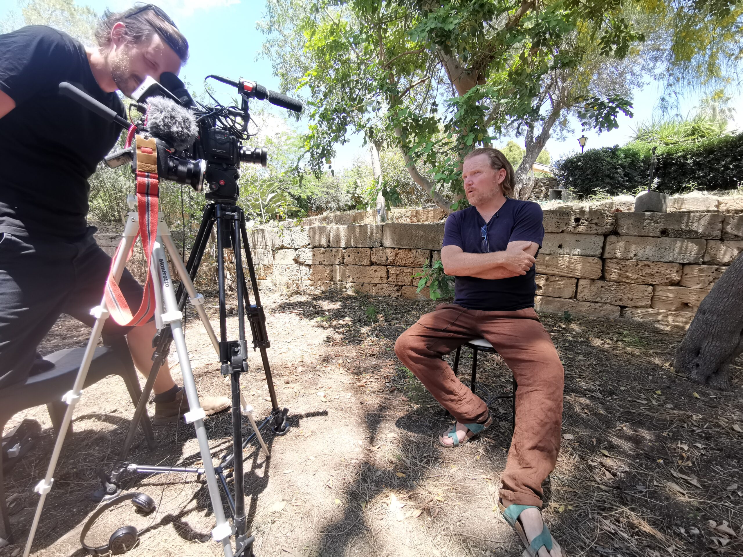 A cameraman filming a farmer sitting before a wall under a tree. 